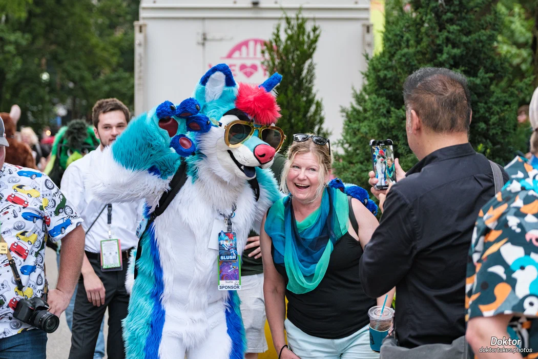 A smiling convention guest posing with colorful fursuiters while another attendee takes a photograph